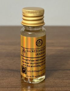 A small glass bottle of ‘Sacred Anointing Oil (Kadosh)’ with gold caps and holographic gold labels standing on a wooden surface. The labels display branding, usage instructions, and contact information.”