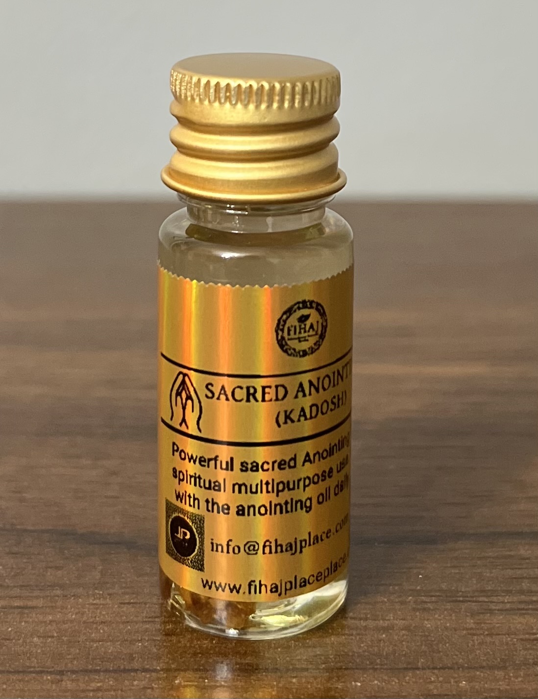 A small glass bottle of ‘Sacred Anointing Oil (Kadosh)’ with gold caps and holographic gold labels standing on a wooden surface. The labels display branding, usage instructions, and contact information.”
